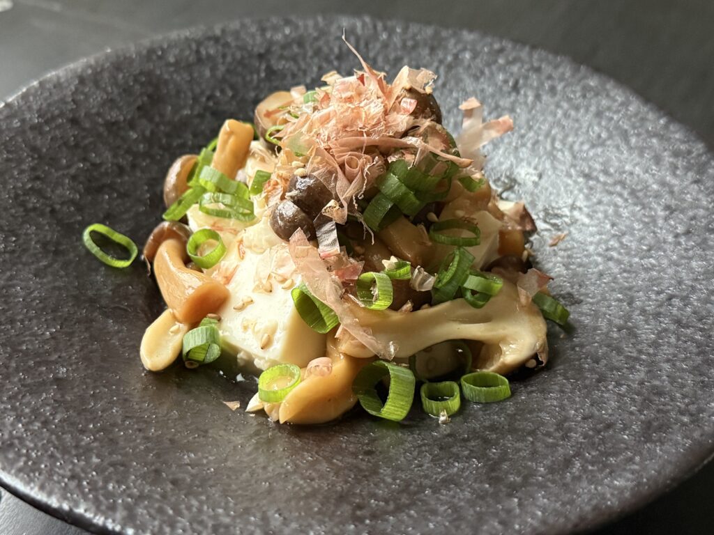 黒い器に盛り付けられた、豆腐としめじを使ったサラダ。鰹節と白ごま、青ネギの色どりが美しいサラダ。