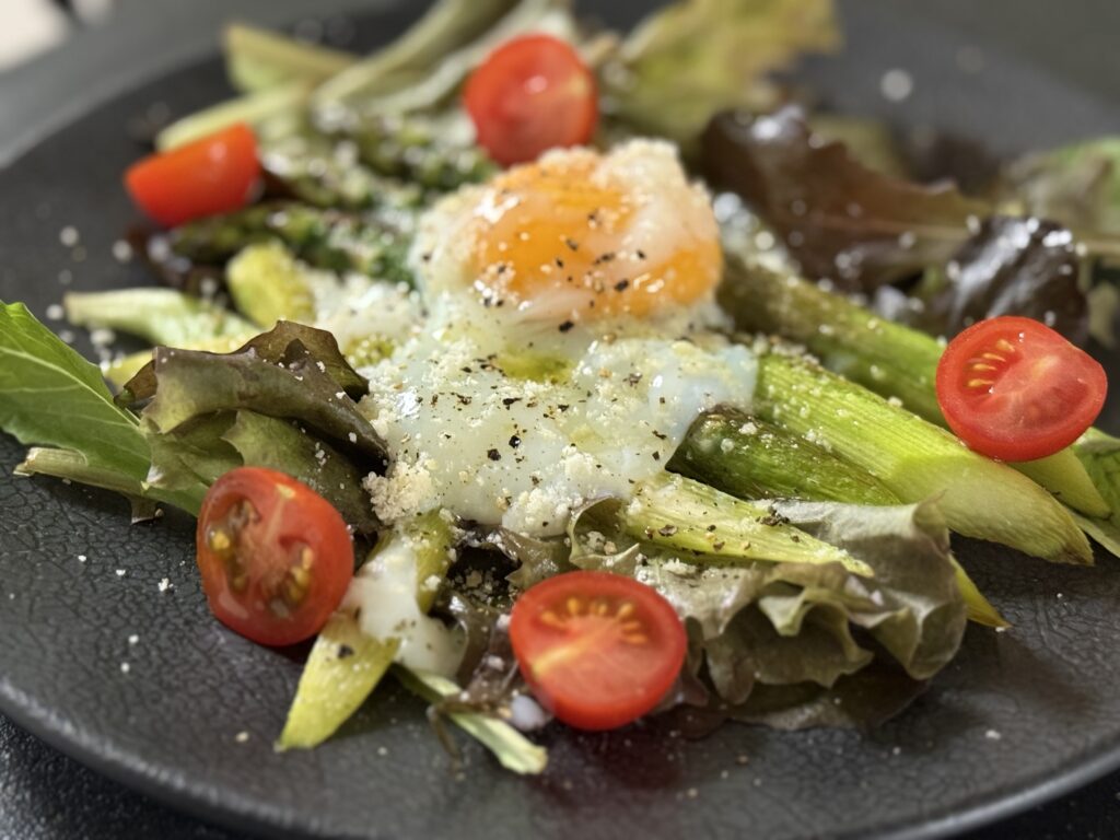 黒い皿に盛り付けられた焼きアスパラと温泉卵のサラダ。ベビーリーフとミニトマトを添え、粉チーズと黒胡椒が全体に振りかけられている。