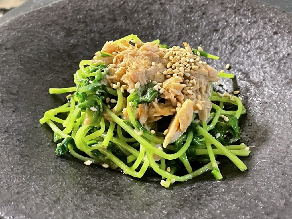 黒い皿に盛り付けられた豆苗とツナの和風サラダ。白ごまがトッピングされている。
