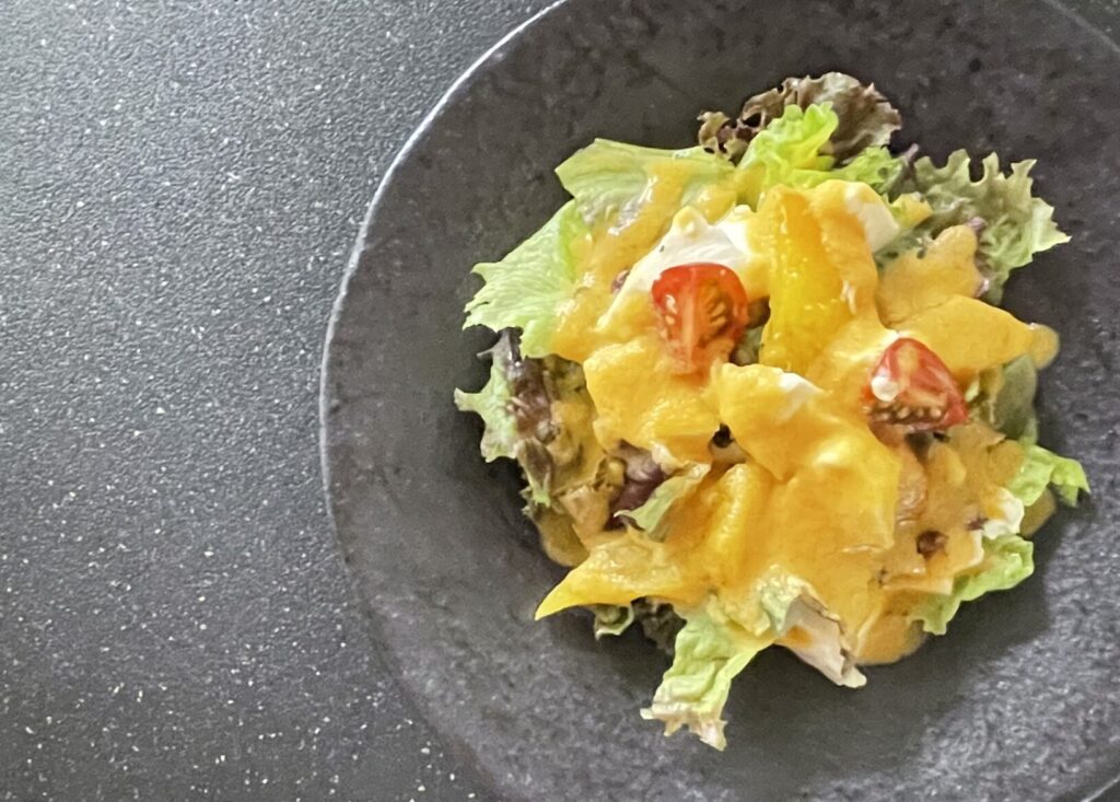 黒いプレートに盛りつけられた、黄色パプリカと豆腐のサラダ。リーフ野菜とミニトマトの上に、鮮やかなオレンジ色のドレッシングがかかっている。