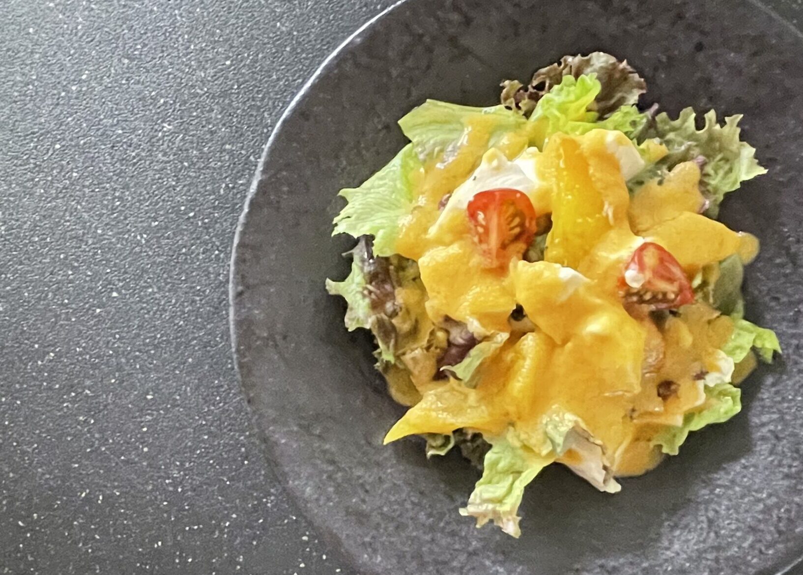 黒いプレートに盛りつけられた、黄色パプリカと豆腐のサラダ。リーフ野菜とミニトマトの上に、鮮やかなオレンジ色のドレッシングがかかっている。