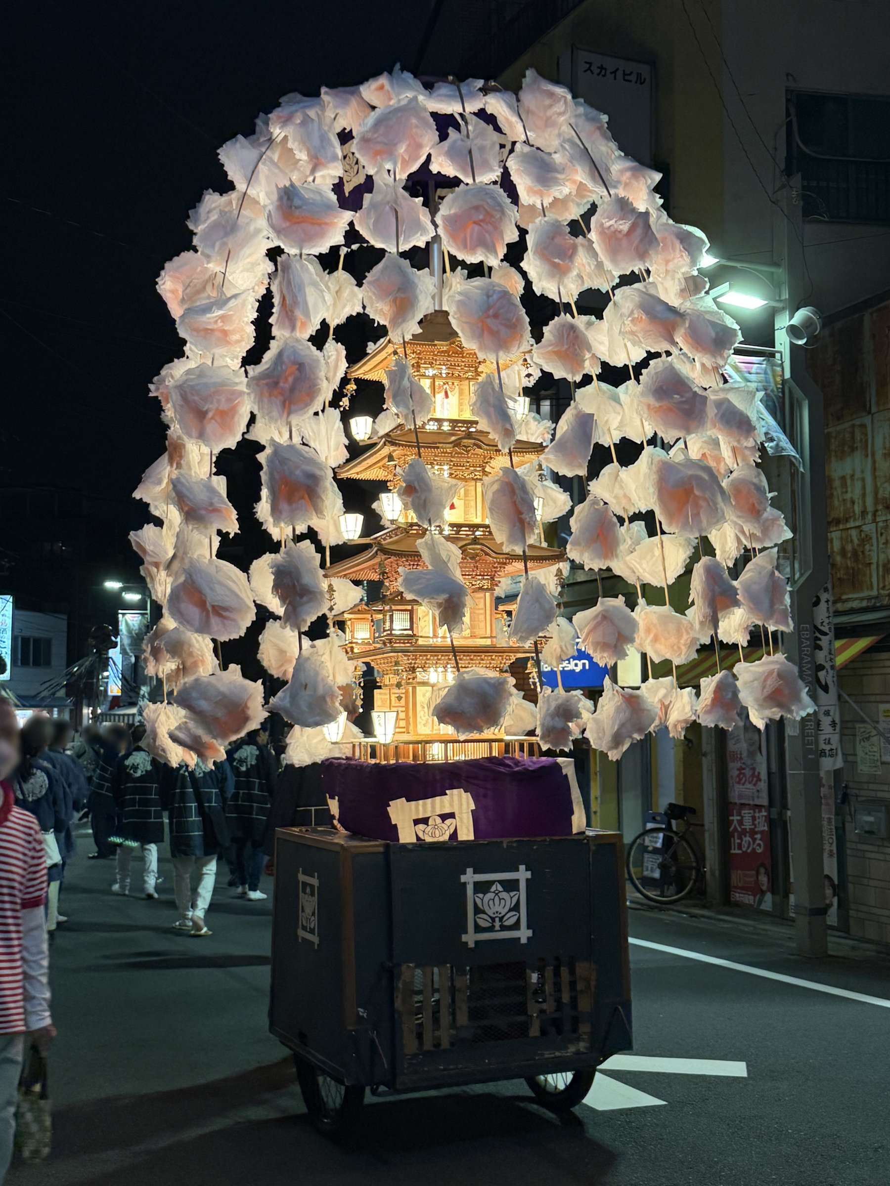 商店街の灯りの中を進む、和紙の桜で飾られた五重塔型の万灯